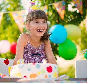 Little girl laughing at a birthday party, sitting in front of a cake and wearing a birthday hat, with balloons in background